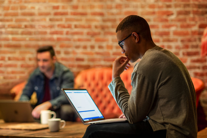 A man sitting at a desk, focused on his laptop computer, with a neutral background.