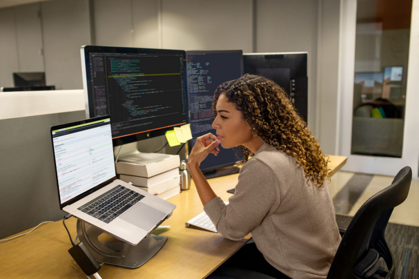 A woman sitting at a desk, working on a laptop with a monitor beside her.