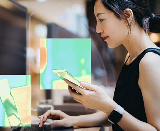 Person sitting at a desk using a smartphone and working on a computer, with heat map highlights over the phone and computer screen indicating areas of interaction.