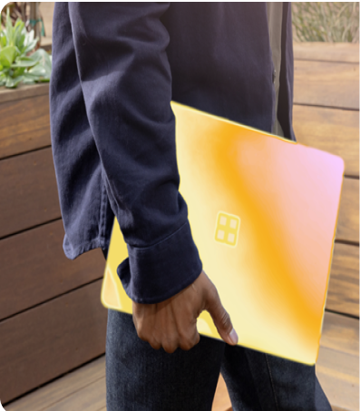 Person holding a yellow laptop with a four-square logo, wearing a dark long-sleeve shirt and jeans, standing in front of wooden panels with greenery in the background.