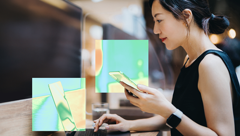 Person sitting at a desk using a smartphone and working on a computer, with heat map highlights over the phone and computer screen indicating areas of interaction.