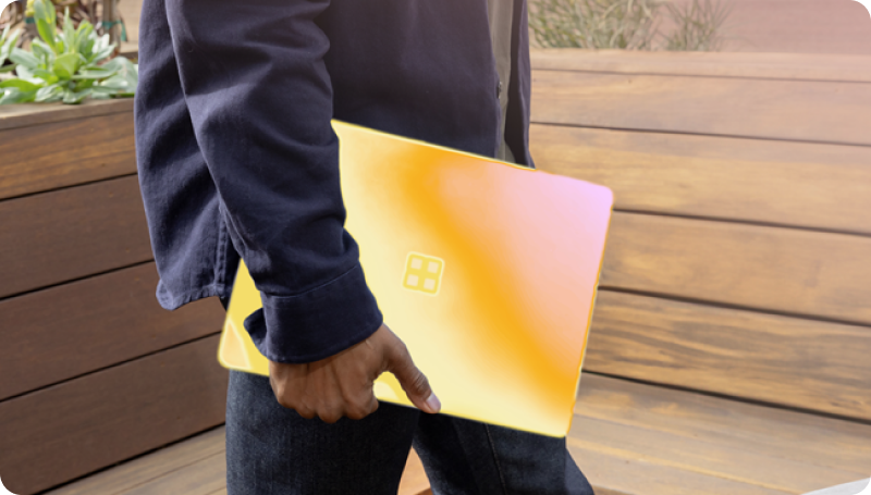 Person holding a yellow laptop with a four-square logo, wearing a dark long-sleeve shirt and jeans, standing in front of wooden panels with greenery in the background.