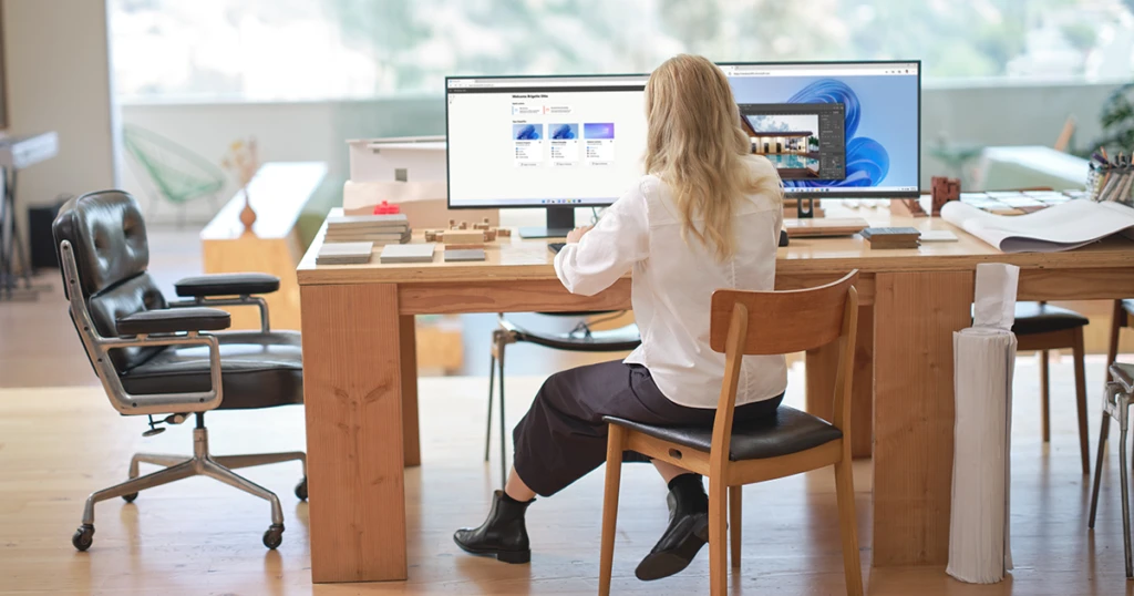 mujer de espaldas, sentada frente a su escritorio, el cual tienen dos pantallas. en las cuales ella esta trabajando.
