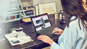Femme travaillant sur un ordinateur portable, assise à son bureau dans son bureau à domicile. Elle a un presse-papiers, du courrier et d’autres fournitures sur son bureau et une étagère en arrière-plan.