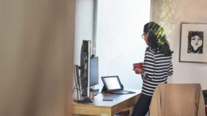 Femme travaillant depuis son bureau à domicile sur un appareil Surface Pro, debout à son bureau avec une tasse dans les mains.