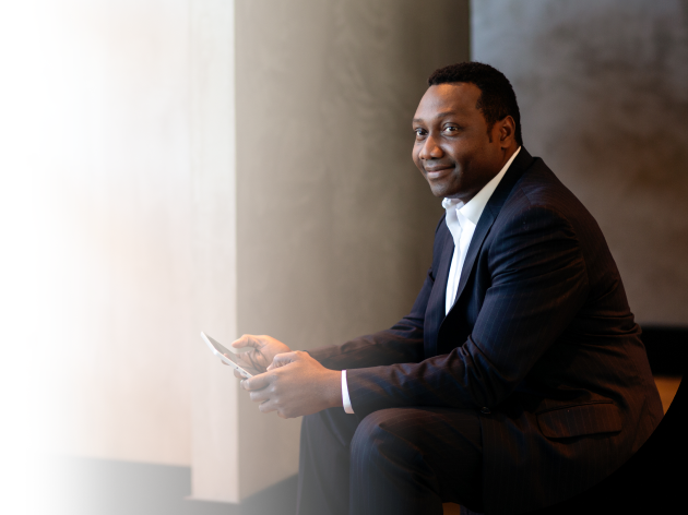 Seorang laki-laki yang berpakaian formal, sedang duduk dan tersenyum sambil memegang tabletnya