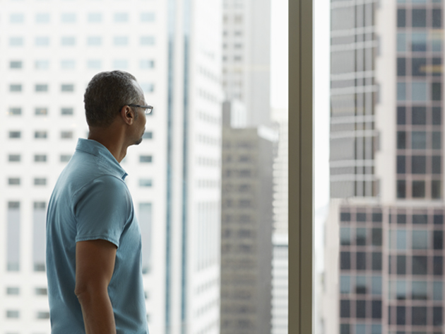 Seorang laki-laki dalam ruangan yang sedang berdiri mentap keluar jendela sambil membelakangi kamera. 
