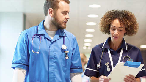 Seorang dokter dan perawat sedang jalan beriringan sambil mendiskusikan perihal pasien mereka. Perawat tersebut memegang sebuah kertas. 