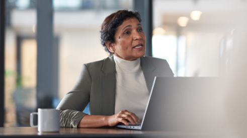 Seorang wanita berambut pendek sedang duduk di meja bersama laptopnya dan tampak sedang mengahdiri sebuah rapat. 