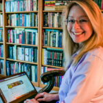 Gina Dyer sits in a library and smiles at the camera while holding her laptop.