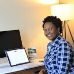 Nyameke sits in front of her laptop and monitor while smiling at the camera.