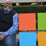 Dwight Jones sits next to the Microsoft sign on the edge of the Microsoft campus. He is smiling.