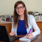 Tait, wearing glasses and a white sweater, sits at a wood desk with her laptop open and a notepad at the ready.