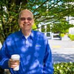 Apple holds a cup of tea as he smiles standing in front of a tree outside a Microsoft office.