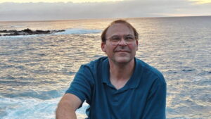 Johnson poses outside in front of an ocean view; he is smiling towards the camera in a navy-blue polo shirt.