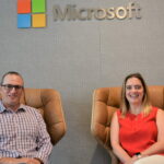 Yedinak and Laves sit and smile below a Microsoft logo inside a Microsoft building.