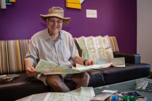 Apple smiles as he reads an unfolded traditional map. He’s wearing an explorer’s hat and shorts and a short-sleeved shirt.