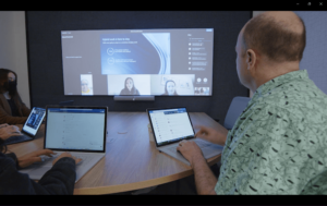 Microsoft employees attend a meeting at a curved table that faces a large wall display showing virtual attendees and meeting content.