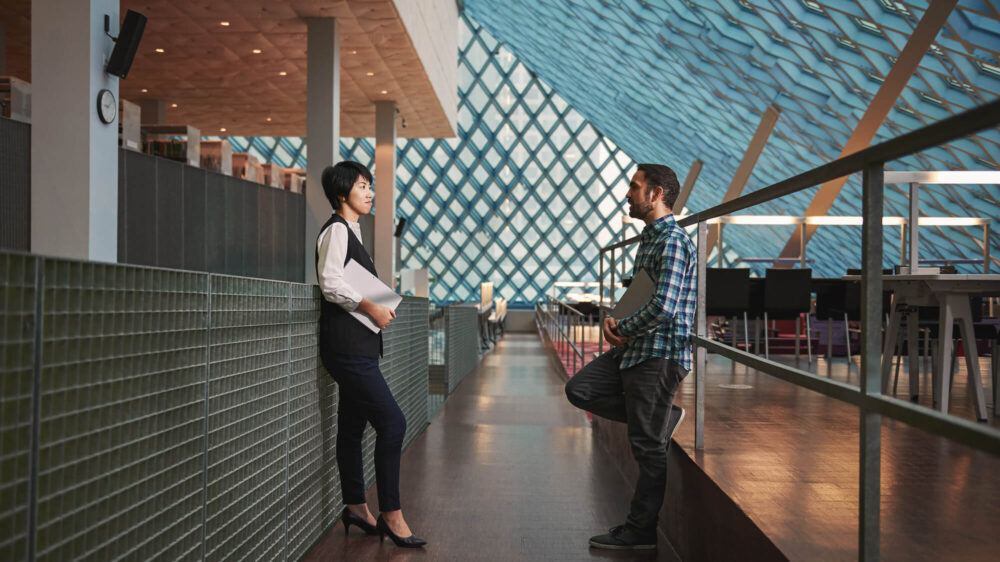 Two people talk together in a passageway in a large open area in a building.