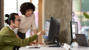 A man and a woman working within Microsoft Teams on dual monitors in an open office setting.