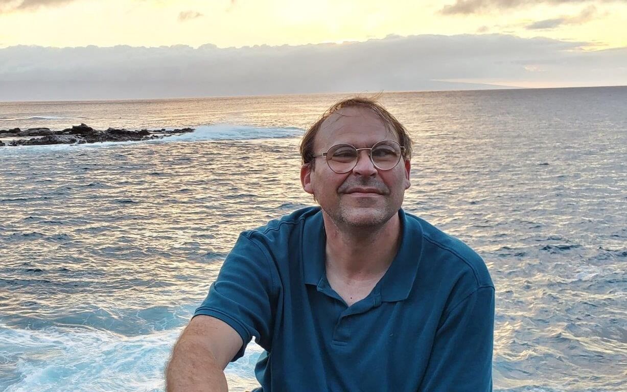 Johnson is outside facing the camera smiling, the sun is setting over the ocean in the background.