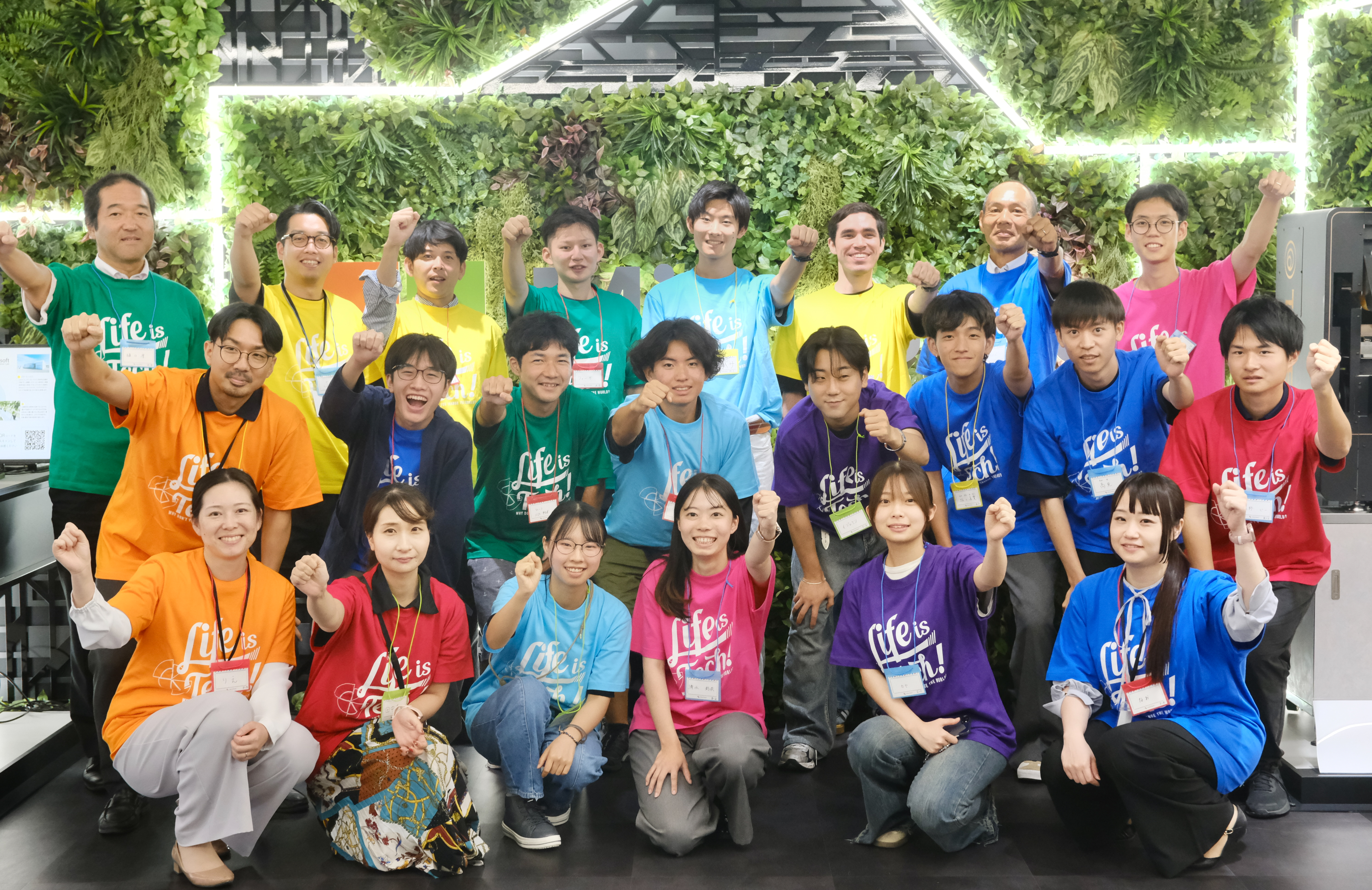 Life is Tech! のカラーTシャツを着たメンバーが観葉植物の壁の前でガッツポーズをする姿。