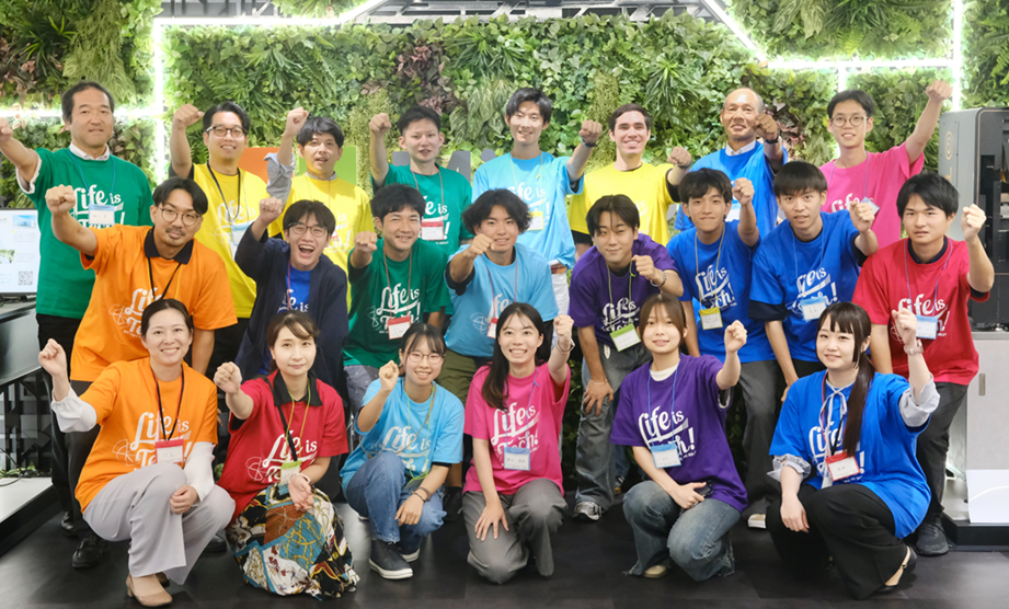  カラフルな「Life is Tech!」Tシャツを着たグループが、緑の植物壁の前で拳を上げてポーズをとっている。