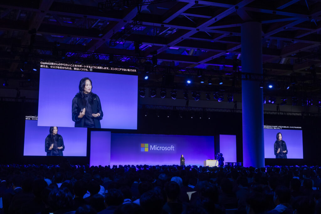 大規模な観客に向けて、Microsoftの基調講演者がステージでプレゼンを行い、大画面に発表内容が映し出されている。
