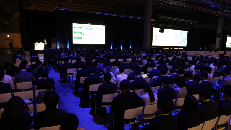 多くの参加者が座って、大画面に映し出されたプレゼンテーションを視聴している会議ホール。