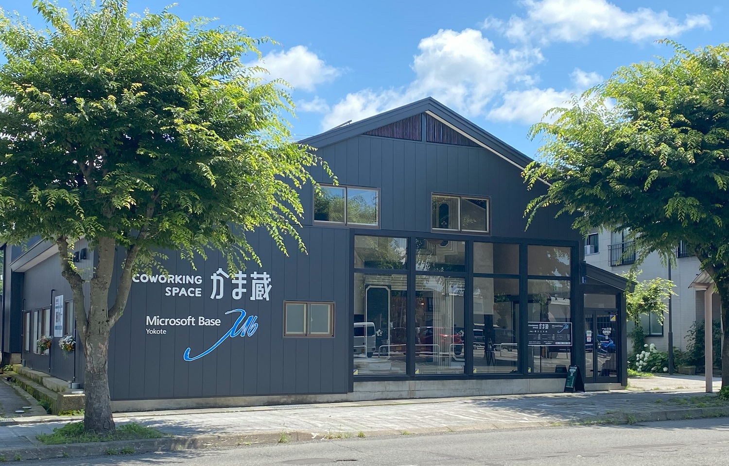 「COWORKING SPACE かま蔵 Microsoft Base Yokote」と外壁に書かれた、緑の木々に囲まれたダークグレーのモダンなコワーキングスペースです。