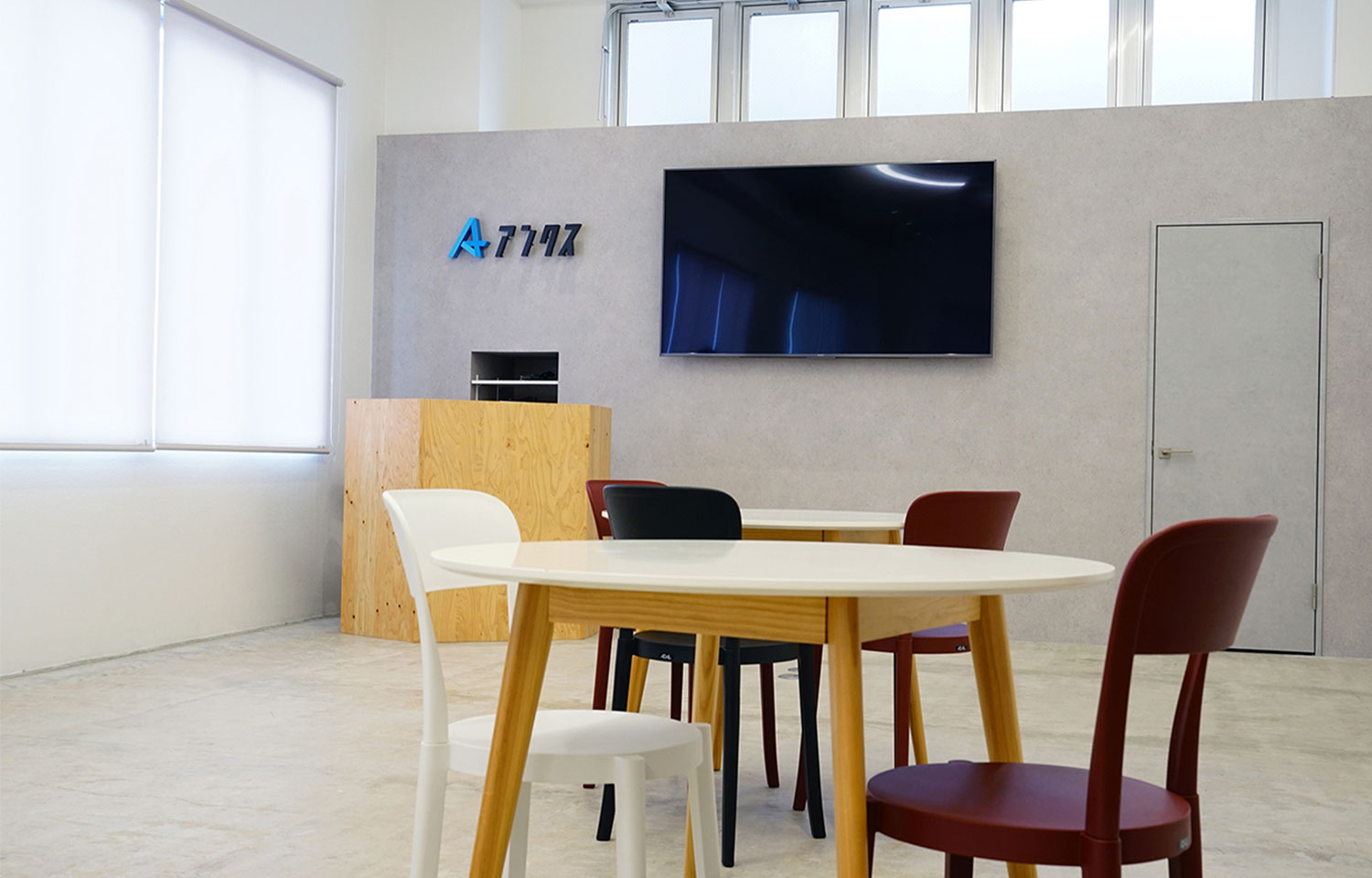 My thought process  清潔でモダンなオフィスまたは待合室。白い丸テーブルと複数色の椅子が手前にあり、奥の壁には「A 7747」のロゴと大型テレビが設置されている。左手には白いブラインドの窓がある。