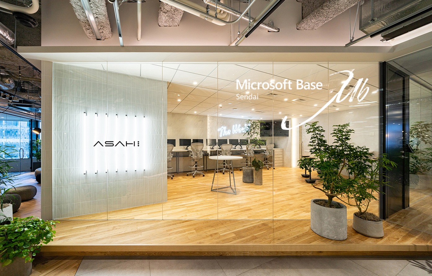  ガラス壁に「Microsoft Base Sendai」と「ASAHI」と表示された、木製フローリングとオフィスチェア、観葉植物のあるモダンなオープンワークスペースです。