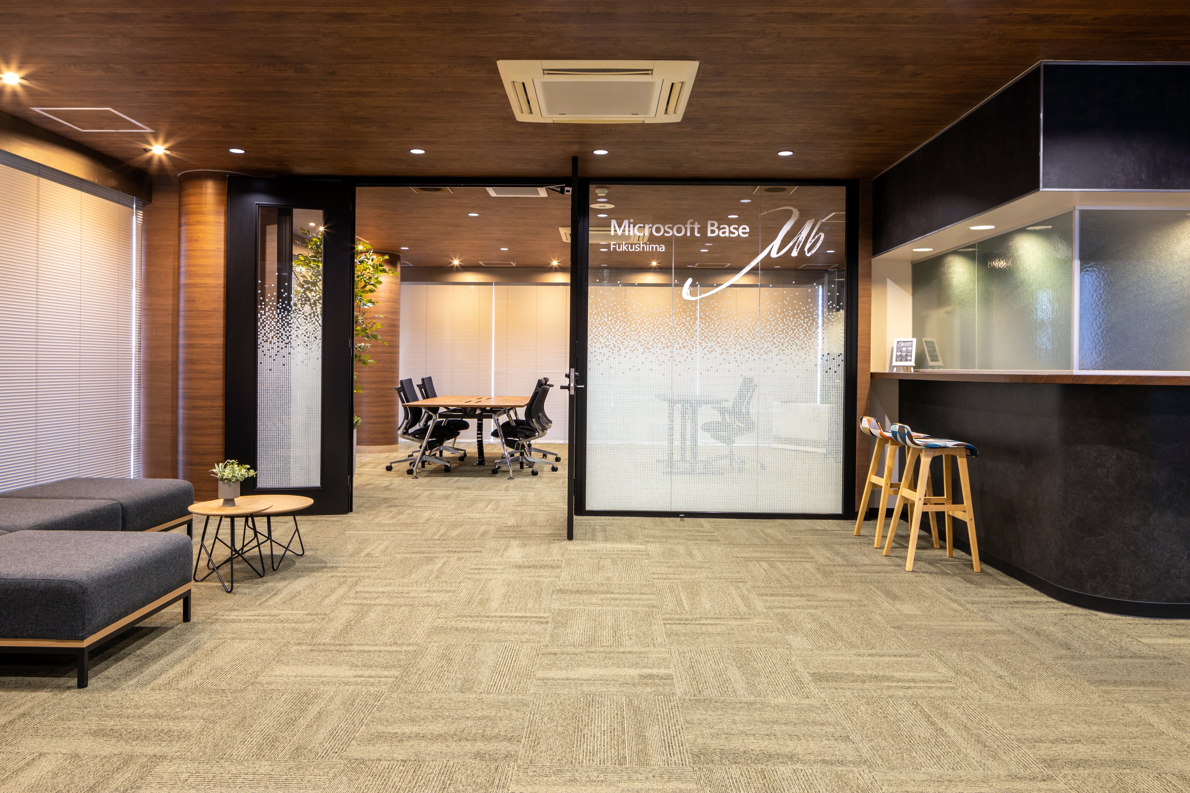 ガラスパーテーションに「Microsoft Base Fukushima」と表示されたスタイリッシュなオフィスロビーで、会議室のオフィスチェア、ラウンジソファ、バースツール、温かみのある木製天井が特徴です。