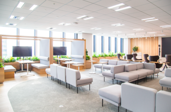 ブース席と独立モニター、薄いグレーのソファ、窓際の植栽がある共同利用ラウンジ。