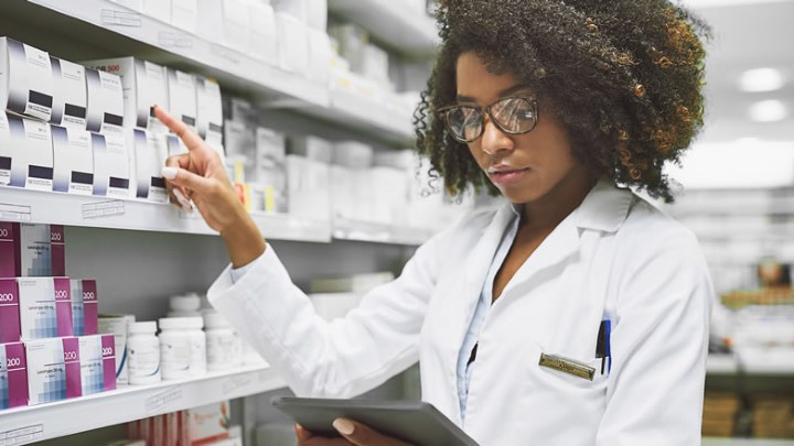 A woman in a pharmacy setting pointing at medications on a shelf and cross-referencing information on a tablet she is holding.