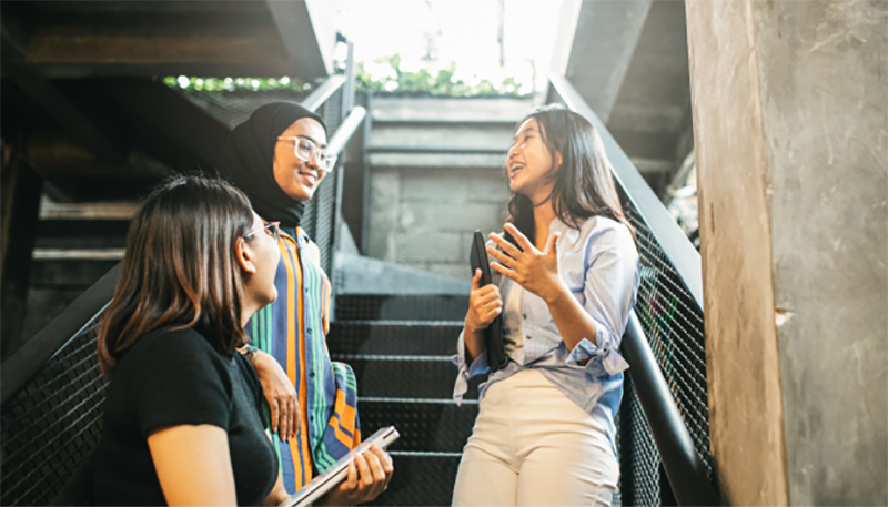 Três mulheres segurando livros conversam em uma escada