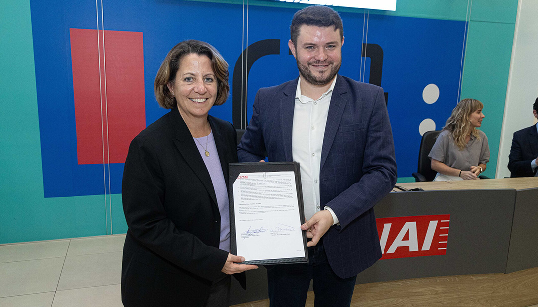 Duas pessoas em um ambiente institucional exibem um documento assinado, com um balcão de atendimento e outras pessoas ao fundo.