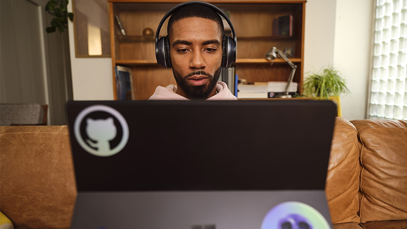 Pessoa usando fones de ouvido trabalha em um laptop em um ambiente doméstico com estante ao fundo