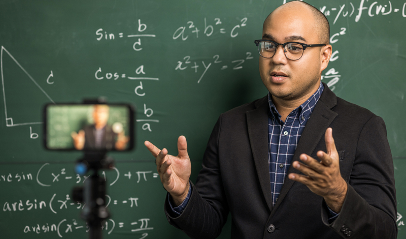 um homem em frente a uma lousa com equações matemáticas falando para um celular em um tripé