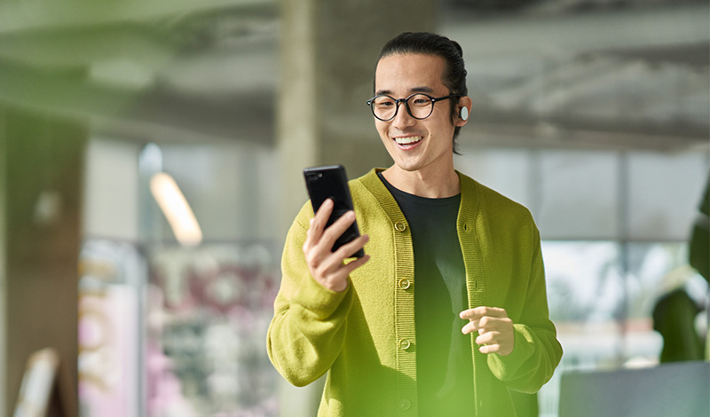 Pessoa em ambiente moderno segurando um smartphone, usando fones de ouvido e vestindo um casaco verde.