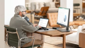 Homem sentado em frente ao computador, ele tem duas telas à sua frente. Em um está sua caixa de entrada do Outlook e em outro, um gráfico e uma tabela no Excel.