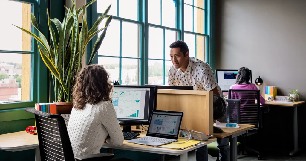 A imagem mostra um escritório, onde uma mulher está trabalhando em seu computador de espadas para nós. Delante dela, um colega fala por cima do divisor de seus lugares. Além deles, outra mulher também está descalça em nós e usa seu laptop.