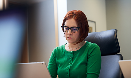 An image of a red-haired woman with a green shirt and glasses, sitting inside an office working on her laptop.