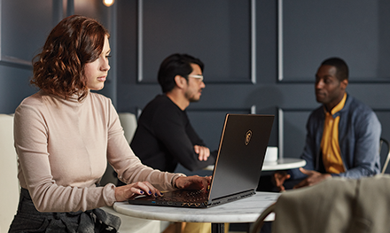 A remote female tech worker on her laptop while two co-workers have a discussion in the backgroud.