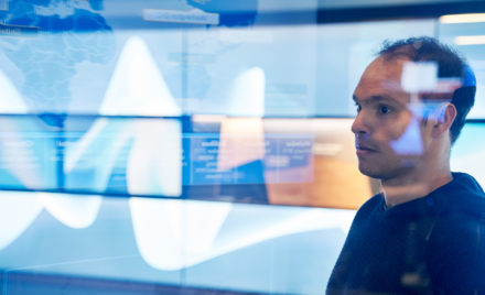Man in hooded sweater/sweatshirt inside a secure room who is looking at data and a geographic area displayed on a large monitor which is behind glass walls with reflections.