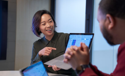 Smiling woman in a meeting room holding up a Surface laptop presenting statistics. Blurred man in foreground.