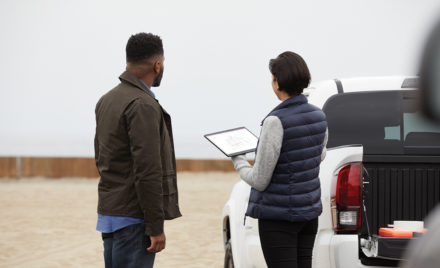 Two adults outside collaborating at a work site while using Microsoft OneNote. Female holding a black Microsoft Surface Pro X in tablet mode with male at her side.
