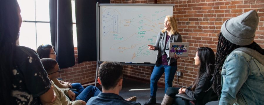 Female developer speaking in front of a white board during team stand up meeting, holding a Surface laptop personalized with stickers.