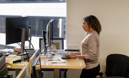 Real people, real offices. Black female developer doing focused work at a standing desk in enterprise workspace.