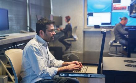 Man in a collared shirt working on a server station inside a secure room. Coworkers and large monitors are in the background.